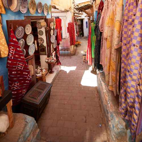 A narrow alleyway in Marrakech flanked by vibrant stalls showcasing textiles, clothing, and handcrafted items.