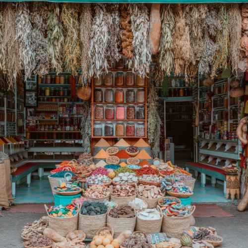 A richly adorned Marrakech spice stall displaying an assortment of spices, herbs, and dried plants, with colorful baskets, ceramic containers, and wooden shelves filled with neatly organized jars in the backdrop.