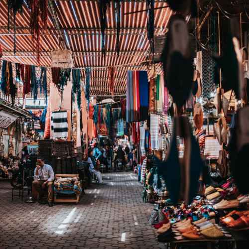 A bustling Marrakech marketplace with rows of stalls showcasing colorful textiles and crafts, under a striped canopy, and traders and shoppers engaging in commerce.